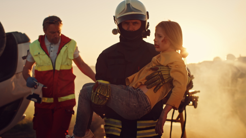 Brave Firefighter Carries Injured Young Girl to Safety. In the Background Car Crash Traffice Accident with Courageous Paramedics and Firemen Save Lifes, Fight Fire
