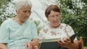 Two elderly women view old photos in album outdoors. Women talking, smiling while sitting on a bench in a green garden. Slow motion 4k video. Closeup.