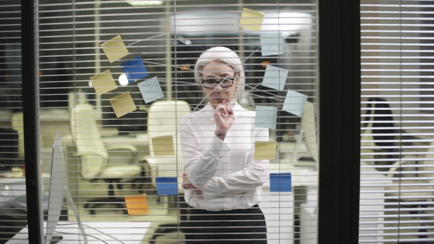 Thigh-up shot of formally dressed Caucasian female employee with grey hair and glasses standing in front of glass partition and working at structural diagram, drawing marker lines between sticky notes