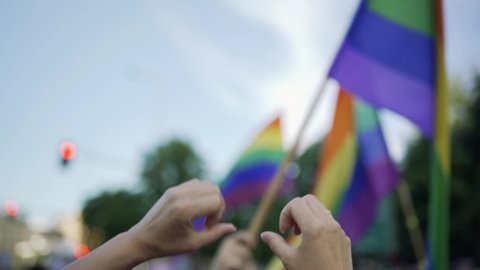 Supporting hands make heart sign and wave in front of a rainbow flag flying on the sidelines of a summer gay pride parade	