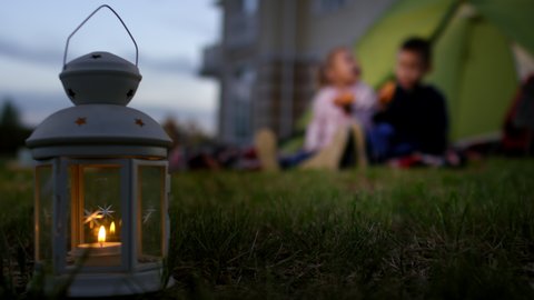 Full rack shot of lantern with burning tealight on grass lawn in twilight, and happy pre-teen Caucasian brother and sister sitting on blanket near camping tent in back yard and eating hotdogs