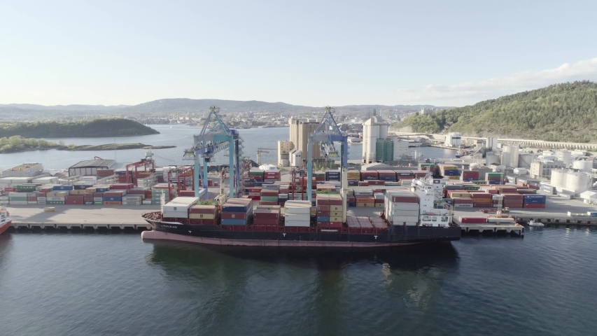 4K aerial with pan/tilt motion of international maritime container port with cranes loading shipping containers on a cargo ship with a smoking pipe in the evening in Oslo, Norway.