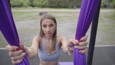 Beautiful girl practicing fly-yoga poses in a hammock outdoor in city park. Aerial, antigravity yoga.