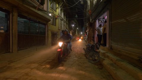 KATHMANDU, NEPAL - SEPTEMBER 2018: SLOW MOTION: Cars and motorbikes drive past local people walking down a dark and dusty street in Kathmandu. Busy third world country city streets on autumn night.