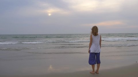 A young slim girl of European appearance with blond short hair, in blue wide trousers Aladdin and white T-shirt stands on the shore with his back to the viewer and looks at the sea in the evening.
