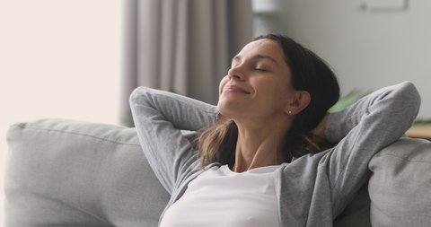 Happy relaxed young woman rest lounge lean on couch enjoy peaceful mood, healthy lazy calm girl dreaming breathing fresh air sit on comfortable sofa in living room on stress free cozy day at home