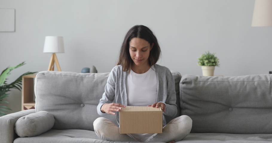 Happy young woman customer sit on sofa open parcel carton box satisfied with online shop order delivery at home, smiling girl consumer unpack package receive good purchase by postal shipping concept | Shutterstock HD Video #1032911129
