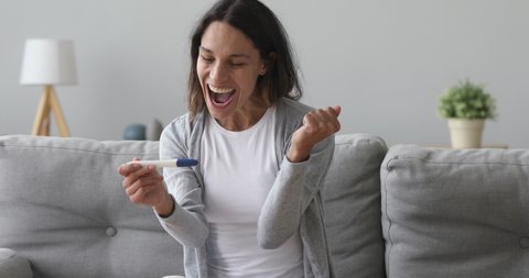 Overjoyed young woman holding pregnancy test stick sit on sofa at home, pregnant lady surprised and happy expecting baby feel excited about positive result on tester after ivf treatment in clinic