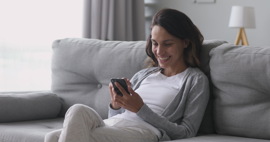 Happy relaxed young woman holding smart phone looking at cellphone screen laughing enjoying using mobile apps for shopping having fun playing games chatting in social media sit on couch at home Royalty-Free Stock Footage #1032911150