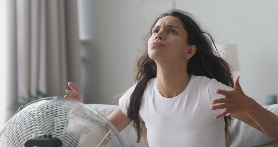 Overheated stressed young woman feel hot suffer from heat high temperature at home sit on sofa in front of ventilator cooling herself with electric fan cooler wind blowing without air conditioner | Shutterstock HD Video #1032911153