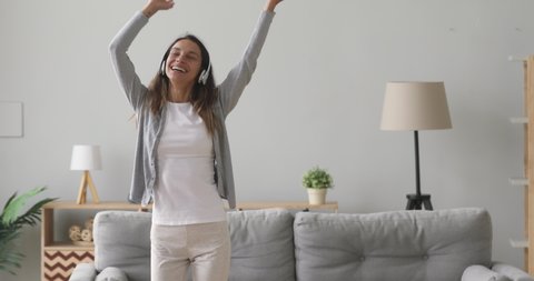 Happy carefree young woman wear wireless headphones dancing alone in modern living room, funny active teen girl jumping moving laughing at home listening to music enjoying new hit song having fun