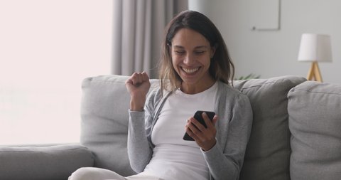 Happy overjoyed girl holding phone celebrate good mobile news surprise bid win game app victory sit on sofa at home, excited young woman winner screaming yes rejoicing success looking at cellphone