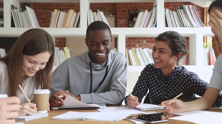 Cheerful multiethnic university college students team laugh at funny joke study together sit at table, happy diverse teen friends group having fun write homework work on project research in library Royalty-Free Stock Footage #1032911252