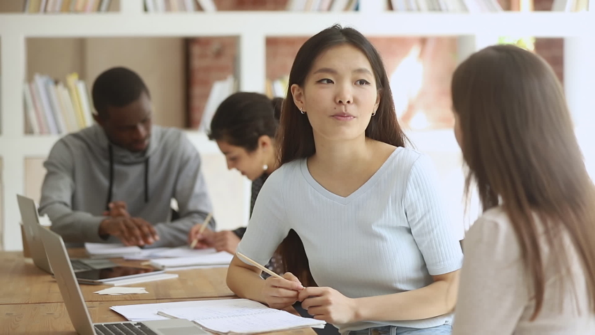 Diverse female college university students talking studying together in classroom, two asian and caucasian teen girls friends discussing high school assignment helping do research project in library Royalty-Free Stock Footage #1032911261