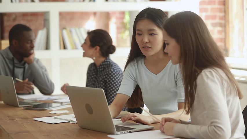 Friendly diverse students talking in classroom using laptop, asian friend or teacher explain teach caucasian classmate girl learn online research course project helping studying together in library Royalty-Free Stock Footage #1032911267
