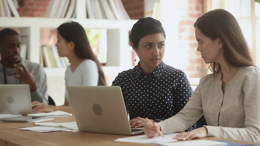 Focused indian young female teacher help caucasian friend student explain computer online project sit at desk in library, mentor teach intern, diverse university team girls learners study together Royalty-Free Stock Footage #1032911285