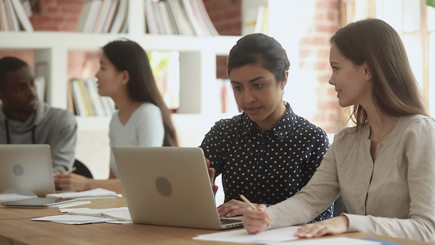 Focused indian young female teacher help caucasian friend student explain computer online project sit at desk in library, mentor teach intern, diverse university team girls learners study together | Shutterstock HD Video #1032911285
