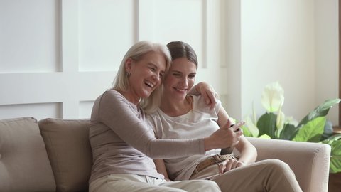 Positive happy mature old mother and grown daughter use funny mobile app take selfie looking at smartphone, smiling aged mom with young woman having fun make phone snapshot self portrait sit on sofa