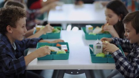 Cute elementary school children sitting by table eating their tasty snacks during lunch in cafeteria at school.