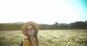 Girl in beautiful summer dress, hat and glasses posing at sunset in daisy field. Slow motion video in 4K.