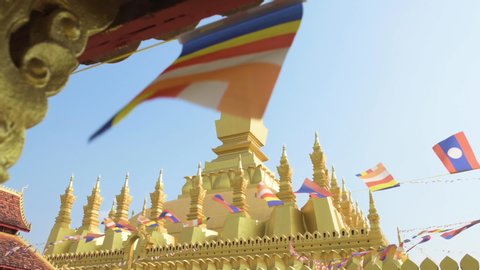 (Selective focus) Slow motion video of Laos flags waving in the foreground with the beautiful Pha That Luang in the background. Pha That Luang is a gold-covered large Buddhist stupa in Vientiane, Laos