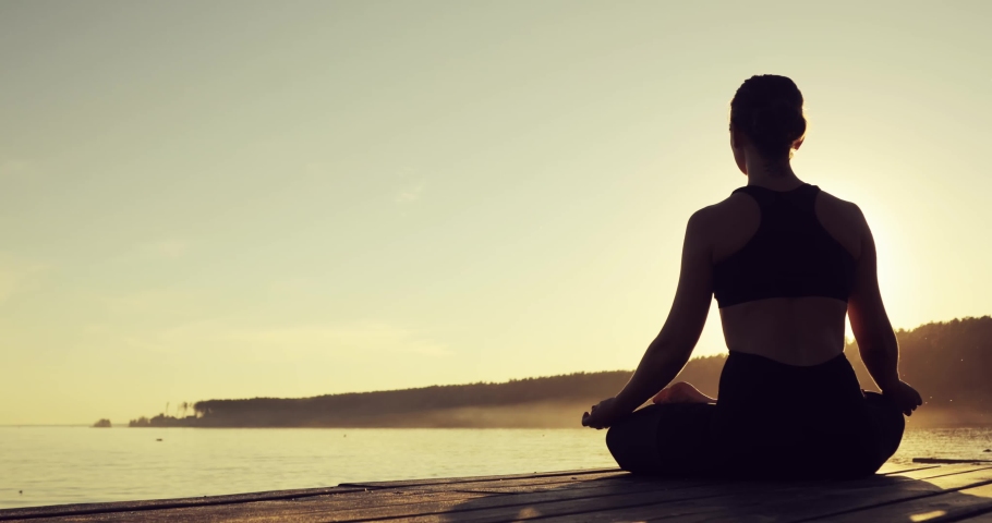 Young woman is meditating in Lotus Pose sitting on pier near the river at sunset. Girl is practicing yoga technique, back view. Silhouette of young slim woman at yellow sunset, low angle shot. | Shutterstock HD Video #1032940472