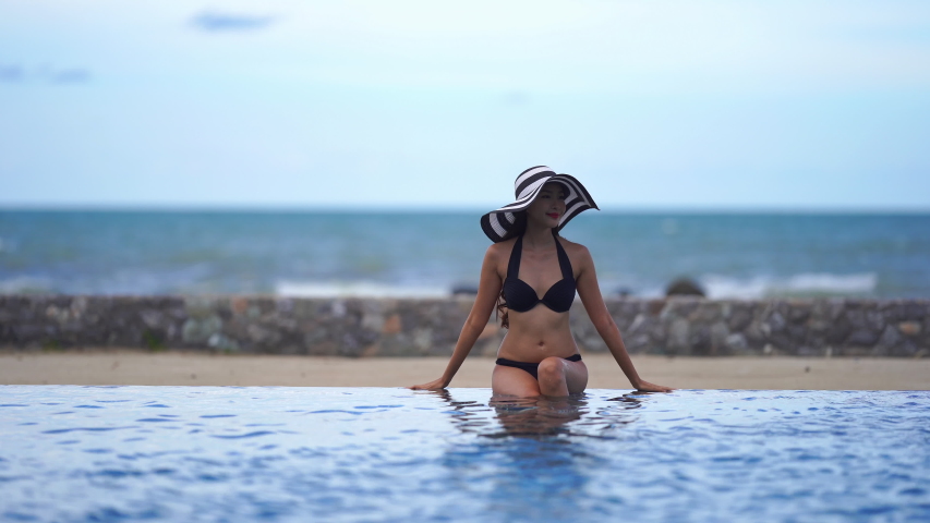 Beautiful smiling woman realxing on the edge of swimming pool wearing a beach straw hat with blurred shore in the background, day, real time, static camera, eye level shot