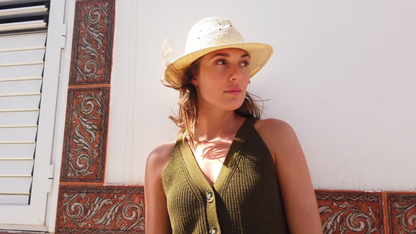 Tilted angled shot of a stunning female model looking sideways, wearing a straw hat and a green knitted top, white wall in her background.
