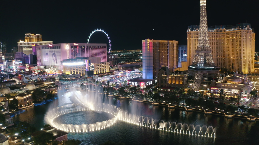 Crazy night full of neon lights, bright twinkles and other nightlife. Fountain show on the background of Eiffel Tower and Ferris Wheel. The Strip. Las Vegas. Nevada. Jul 2019. Aerial 4k.