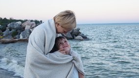Mom hugs her son on the beach and wraps his blanket that would warm. He strokes the boy's face with his hand. They smile sweetly. Mother and son