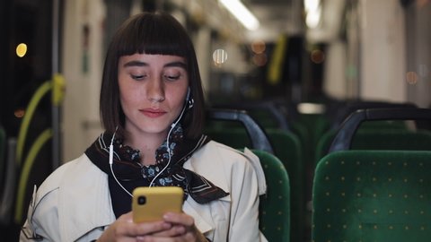 Young stylish woman in headphones listening to music and browsing on mobile phone riding in public transport. City lights background.