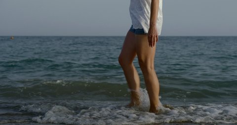 Women's legs in denim short shorts close-up go on a sandy beach near the sea and kick the sea waves.