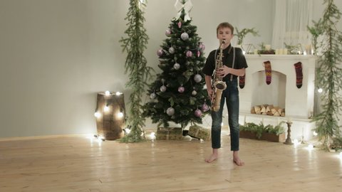 Dolly shot a boy in dark jeans and shirt playing saxophone in a room with Christmas atmosphere, decoration. The concept of the celebration, the new year