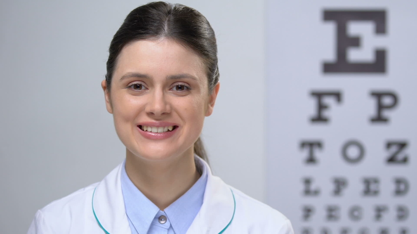 Smiling female ophthalmologist standing against eye chart background, service Royalty-Free Stock Footage #1032951419