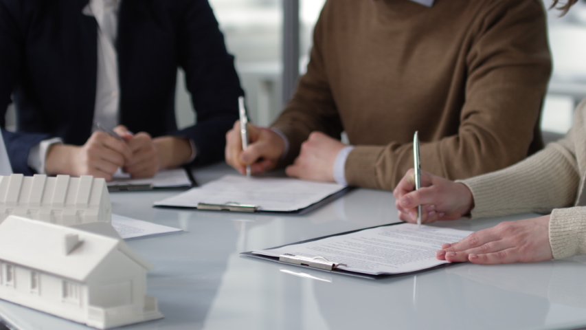 Panning hands shot of man and woman sitting at table with scaled house models in real estate agency and each putting signature on separate copies of legal documents, and professional lady waiting Royalty-Free Stock Footage #1032954146