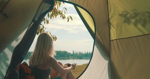Happy traveler woman sitting near tent camp around mountains lake and shoot with her phone camera under sun light enjoying the leisure and freedom. 4K slow motion video