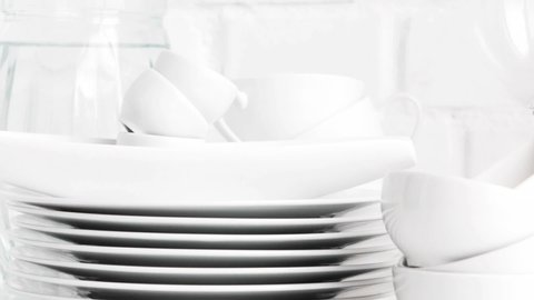 Stacked white porcelain dishes, plates, cups, saucers, mugs in restaurant kitchen interior. Slow pan. White brick wall background.	Selective focus.