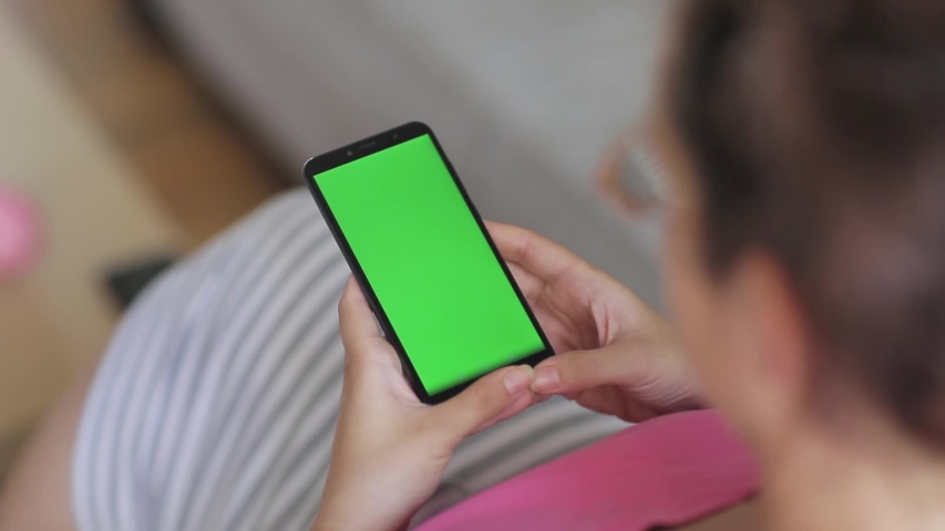 Young woman sitting on a couch at home, using smartphone with green screen, reading social media or watch movie.