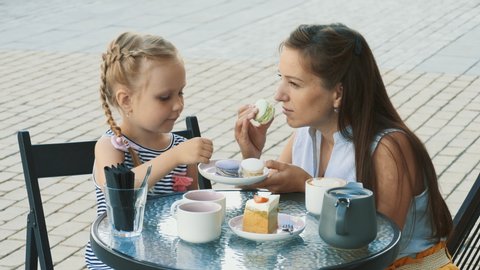 Beautiful brunette mother and her cute little daughter sitting in outdoor summer cafe. Happy family drinks tea with colored macaroons and cake in cafe.