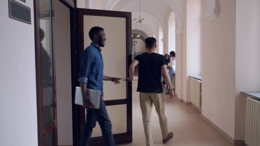 View from back of african male student closing door after lesson and going through corridor in university. Smart boy greeting with friends, talking and laughing. Concept of learning process.