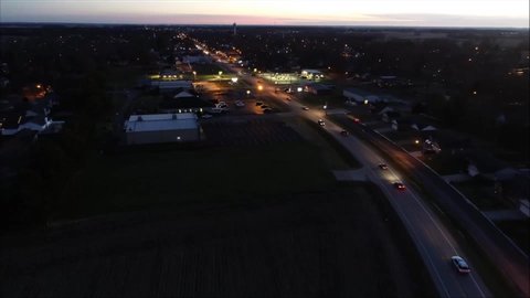 A drone flies over a small town at dusk as cars drive down a two-lane highway. City lights, homes, trees, and a water tower fill the horizon. 