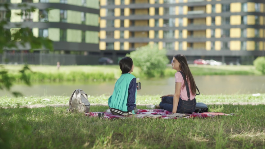 Tracking shot back view of two diverse female students relaxing after classes sitting on lawn near pond and talking. Asian girl drinking coffee, Caucasian pointing at campus sites