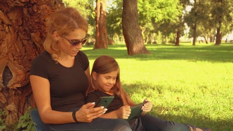 girl and woman are sitting by the tree outdoors. family surfs internet.