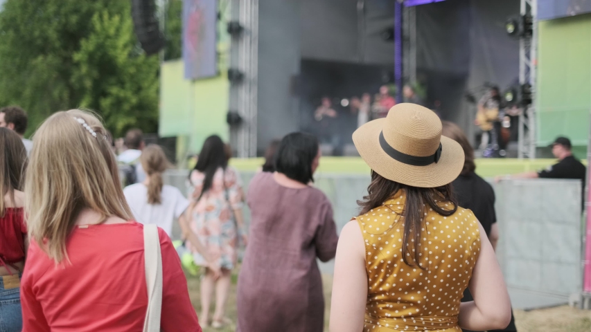 Women are watching concert at open air music festival, back view, stage lights and dancing fan crowd at background