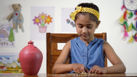 Indian girl inserting coins into a clay money bank (Gulak) - savings concept. Little beautiful girl saving money in a piggy bank - kids financial concept