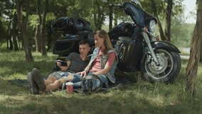 Positive young couple streaming video online on cellphone and chatting while enjoying outdoor leisure near motorcycle. Cheerful friends browsing social media content on smart phone during moto trip.