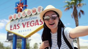 asian female travel blogger recording summer trip to america video by herself in selfie way. young girl doing coming gesture standing on sunny sky with welcome to fabulous las vegas sign background
