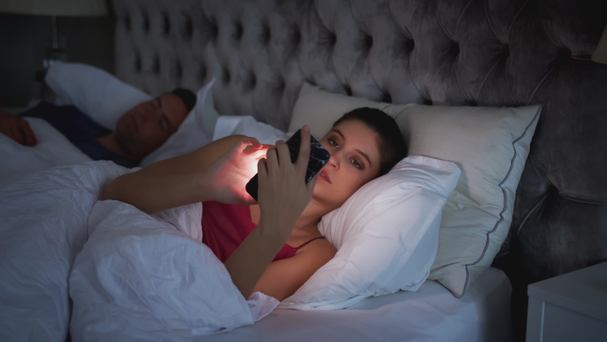 Woman Lying In Bed Checking Mobile Phone Whilst Man Sleeps Next To Her