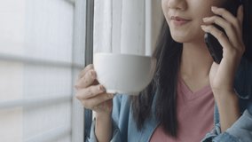 Close up attractive portrait smiling young asian woman drinking coffee and talking on Phone with friends standing beside window at home office.