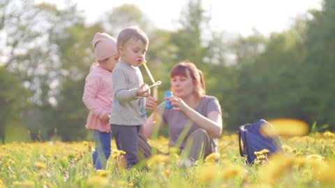 young mother is playing with her children in the city park on a bright sunny spring day. Small son and cute daughter are trying to blow soap bubbles and woman helps them. Happy parenting concept in